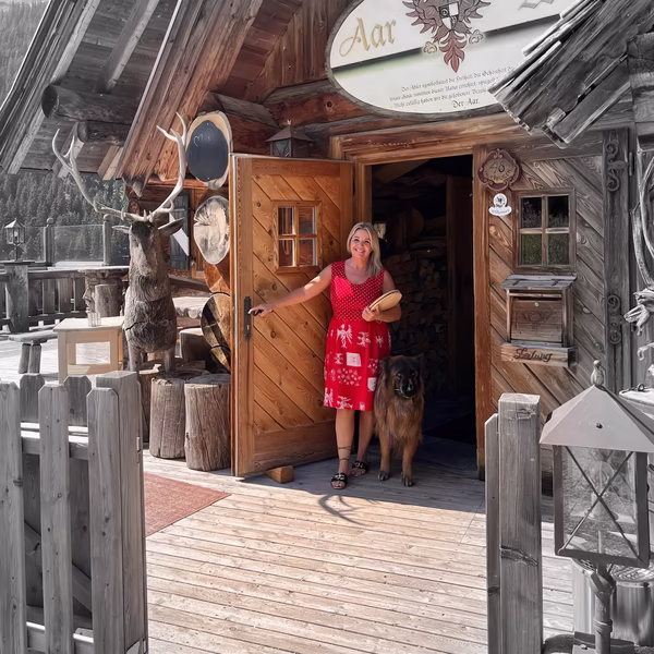 Frau im roten Sommerkleid steht mit einem großen Hund vor dem Eingang einer urigen Holzhütte in den Bergen.