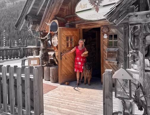 Frau im roten Sommerkleid steht mit einem großen Hund vor dem Eingang einer urigen Holzhütte in den Bergen.