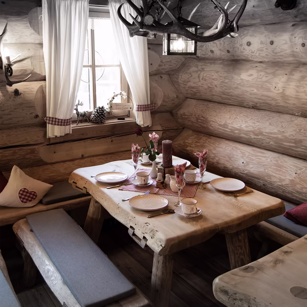 Urig eingerichtete Gaststube mit gedecktem Holztisch, Geweih-Lüster und weißen Vorhängen am Fenster.