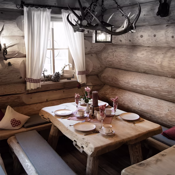 Urig eingerichtete Gaststube mit gedecktem Holztisch, Geweih-Lüster und weißen Vorhängen am Fenster.