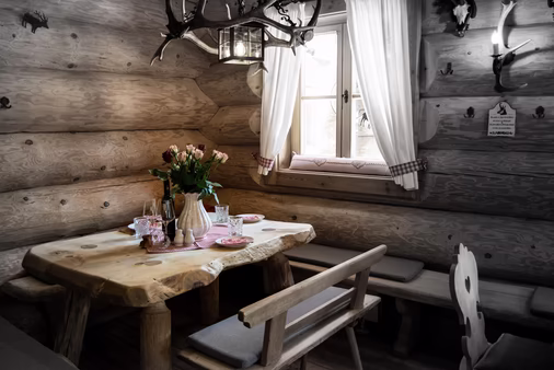 Gemütliche Sitzecke in einer Holzhütte mit rustikalem Tisch, Rosenstrauß und weißen Vorhängen am Fenster.