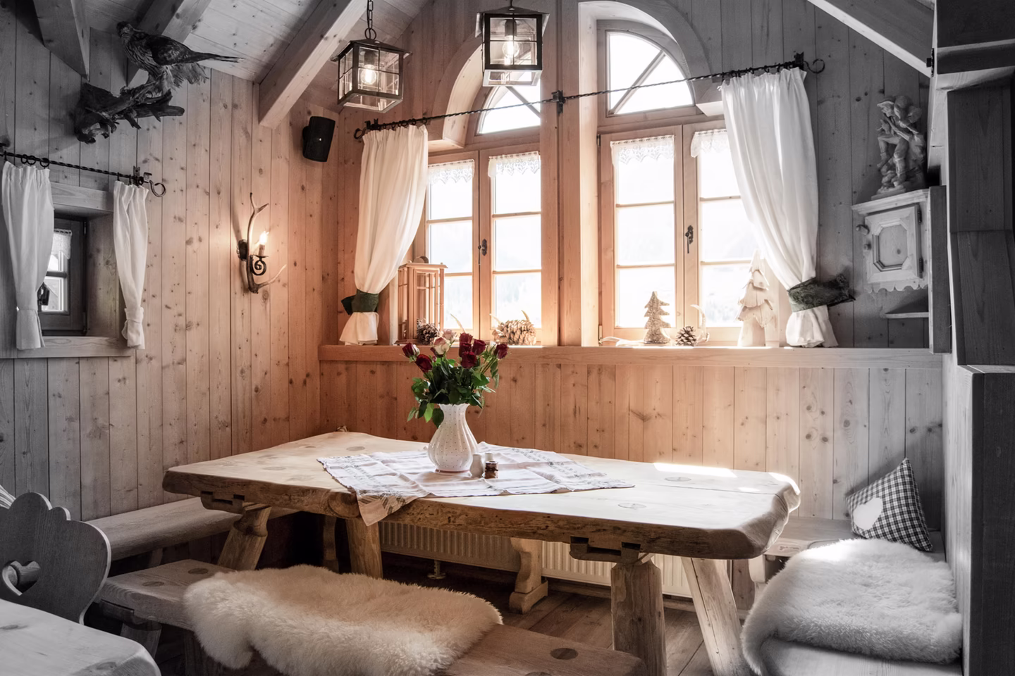 Gemütliche Holzstube mit rustikalem Tisch, Rosenstrauß und Fellauflagen vor sonnigen Fenstern.