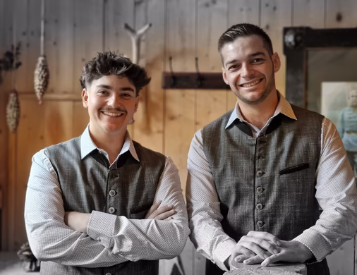 Zwei lächelnde Männer in traditionellen Trachtenwesten stehen nebeneinander vor einer holzvertäfelten Wand.