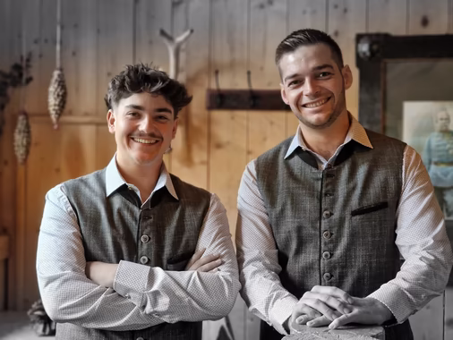 Zwei lächelnde Männer in traditionellen Trachtenwesten stehen nebeneinander vor einer holzvertäfelten Wand.