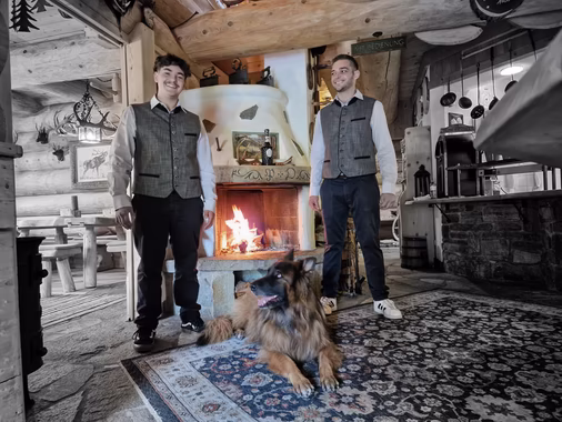 Zwei junge Männer in Trachtenwesten stehen lächelnd am offenen Kaminfeuer einer urigen Holzstube, davor liegt ein Schäferhund auf einem Teppich.