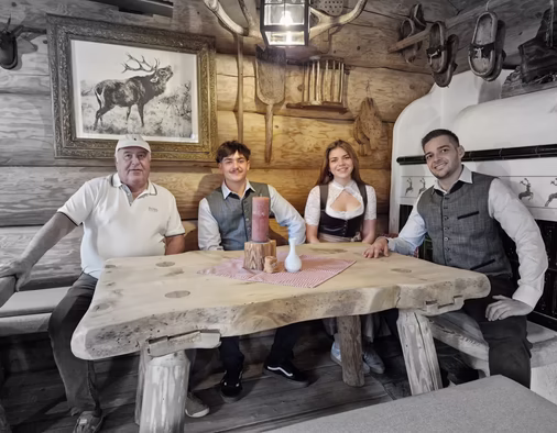 Vier Personen sitzen lächelnd an einem massiven Holztisch in einer gemütlichen Almhütten-Stube mit Hirschbild und Geweihen an der Wand.