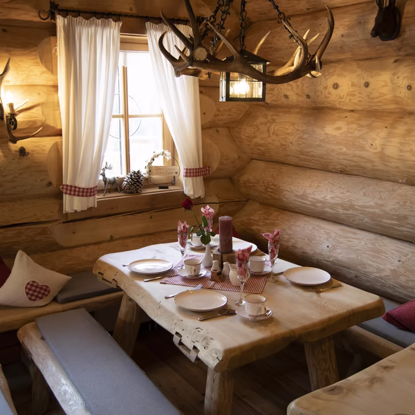 Rustikale Holzstube mit gedecktem Tisch, Geweih-Kronleuchter und karierten Vorhängen am Fenster.
